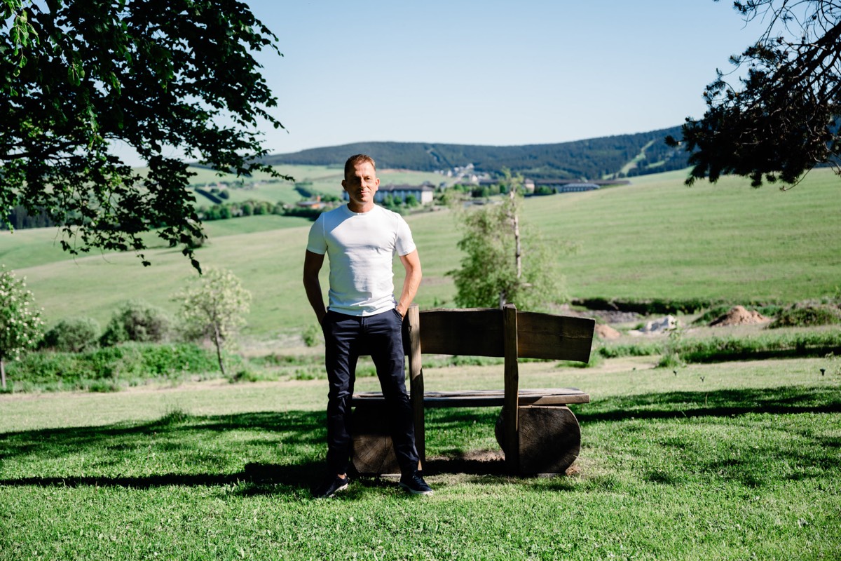 Ralf steht in einer grünen Hügellandschaft neben einer Holzbank